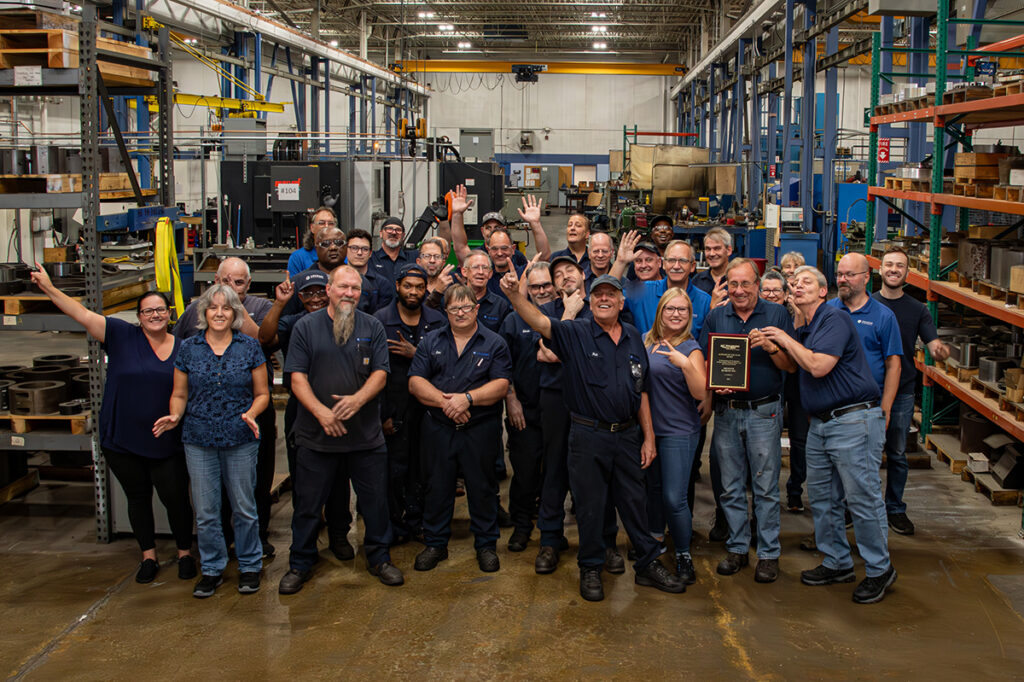 A group of 32 workers from both the shop and the office pose together on the shop floor. Many are smiling, and waving, while others flash #1 gestures as they celebrate receiving the 2025 Supplier of the Year award.